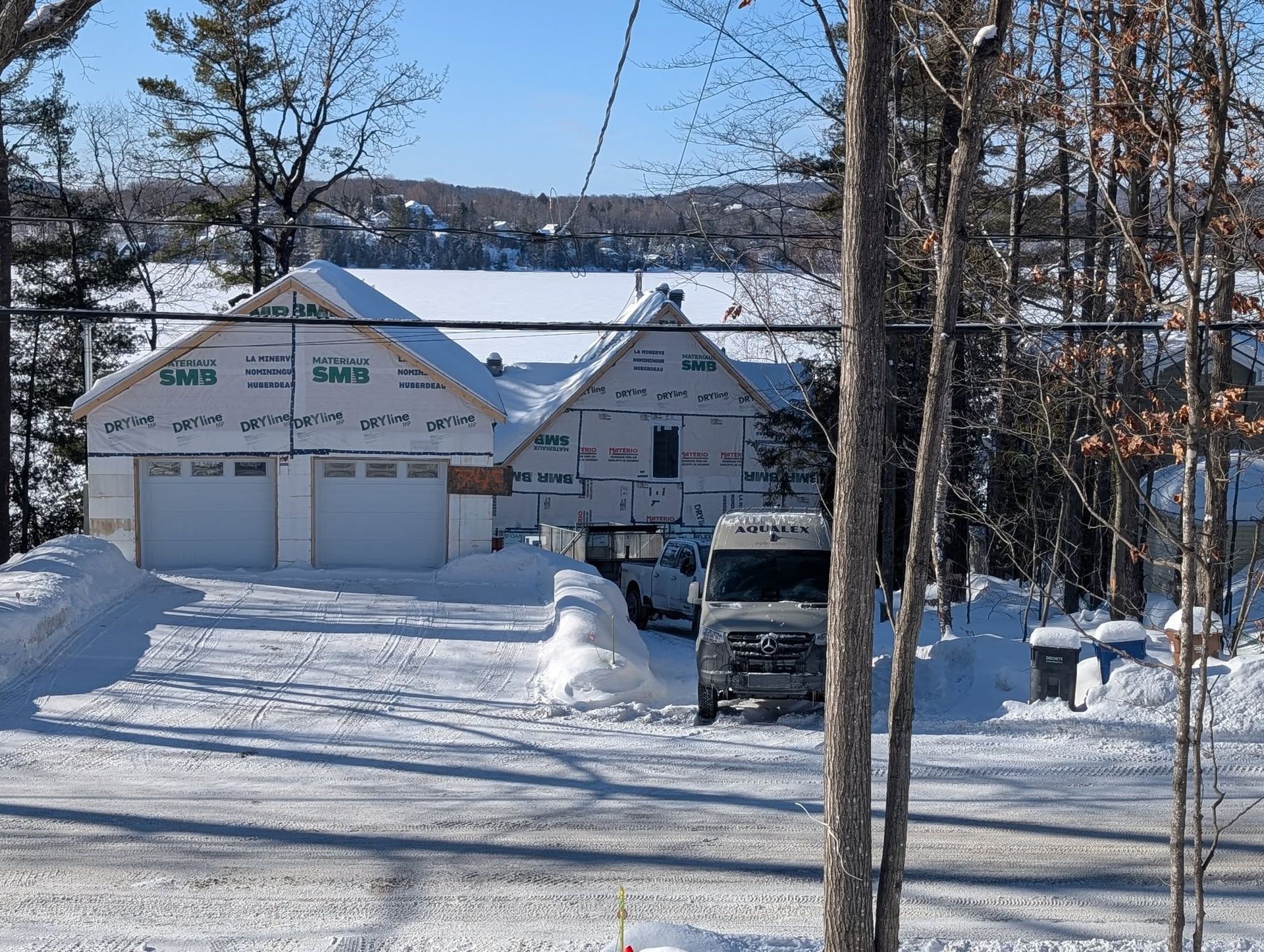 Un garage en construction, recouvert de neige, avec une camionnette garée dans l'allée et un lac en arrière-plan.