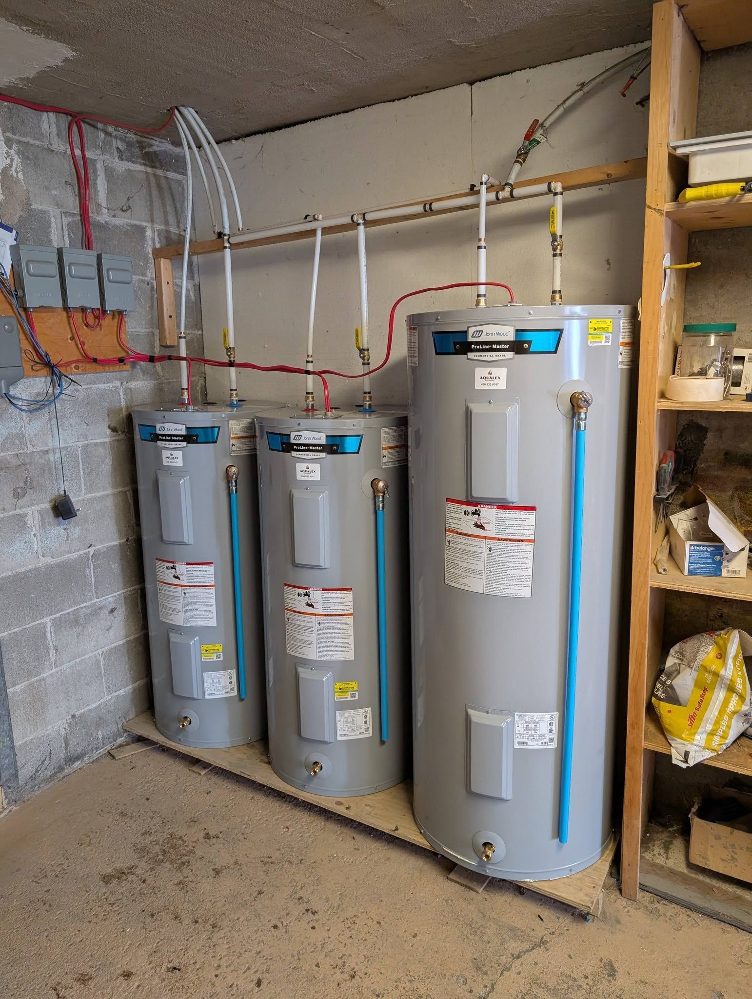 Trois chauffe-eaux grises dans une buanderie. On aperçoit également le câblage électrique et une étagère en bois.