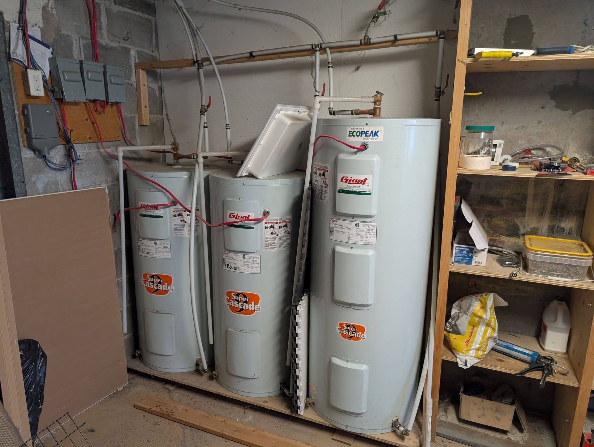 Trois chauffe-eau côte à côte dans un espace de stockage ; étagères en bois à droite, composants électriques à gauche.