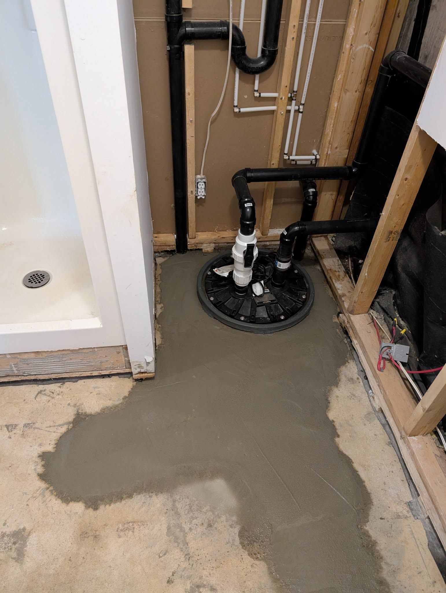 Coin de sous-sol avec douche et pompe de puisard entouré de béton frais. Des tuyaux noirs longent le mur.