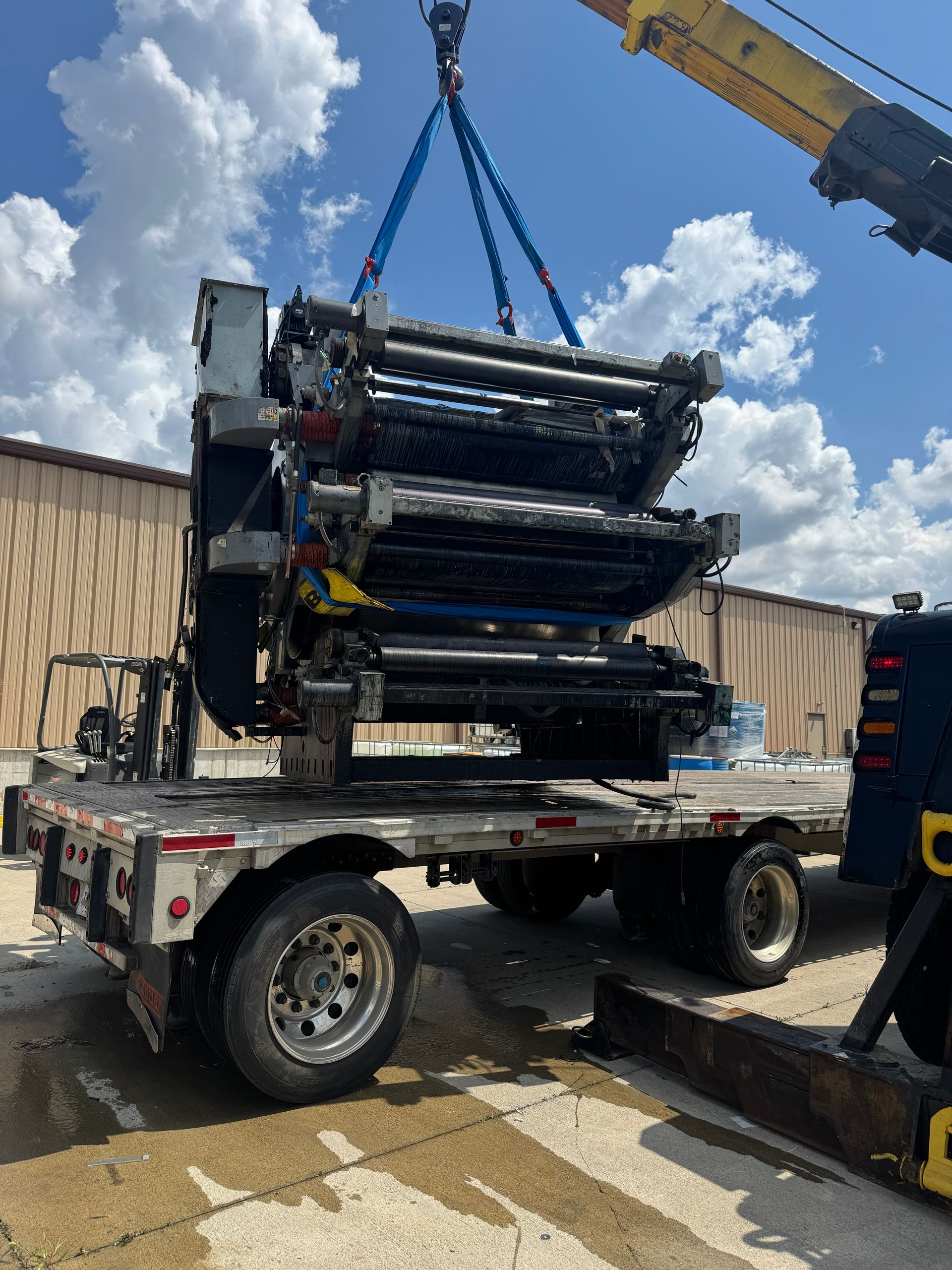 A large printing press module lifted by a crane onto a flatbed truck, outdoors.