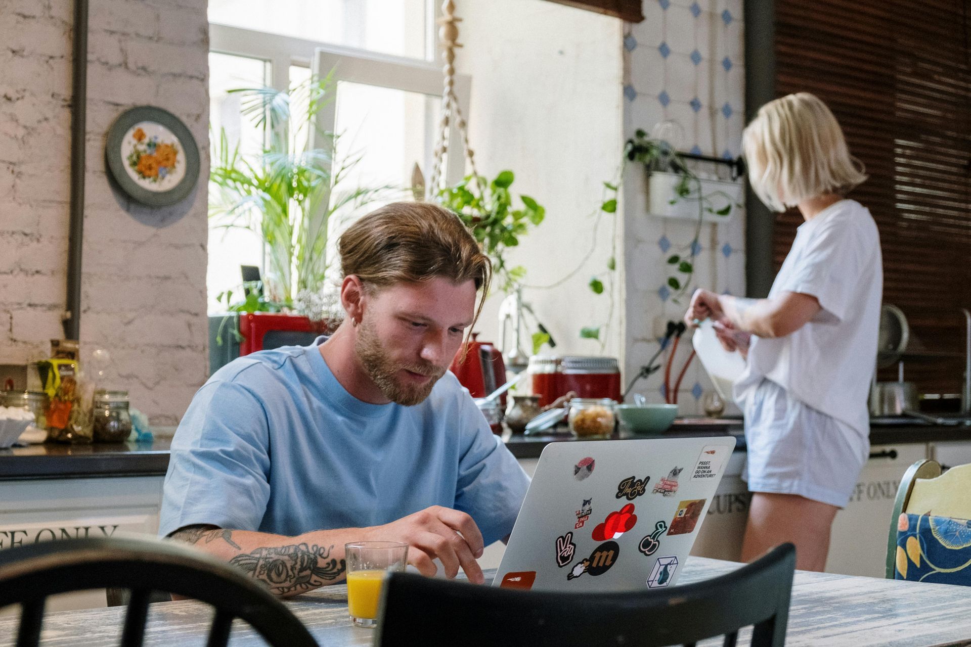 A man is sitting at a table using a laptop computer while a woman stands behind him.