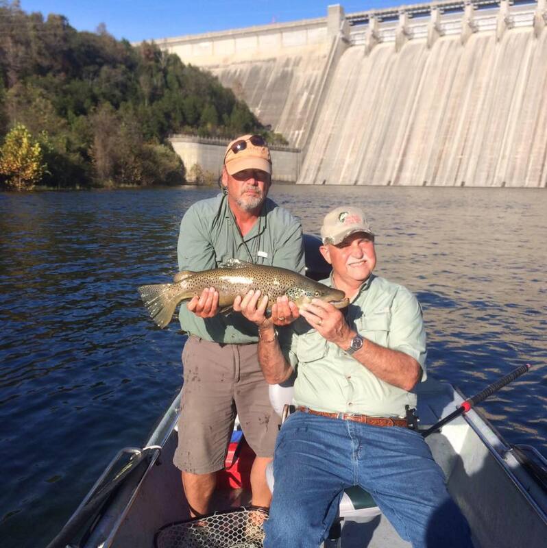 Two men in a boat holding a fish in front of a dam
