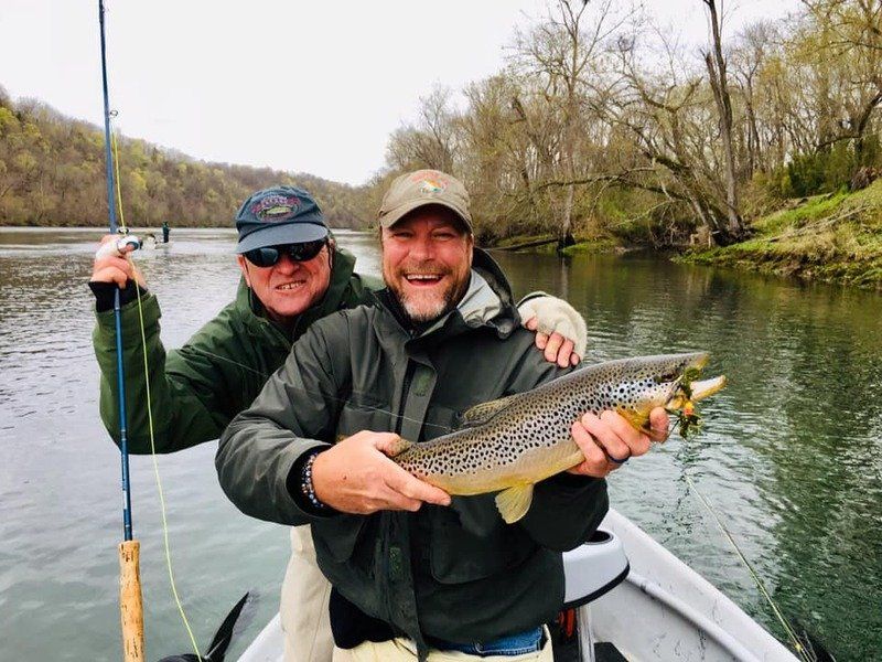 Two men are standing in a boat holding a brown trout.