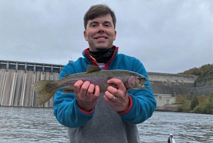 A man is holding a rainbow trout in his hands in front of a dam.