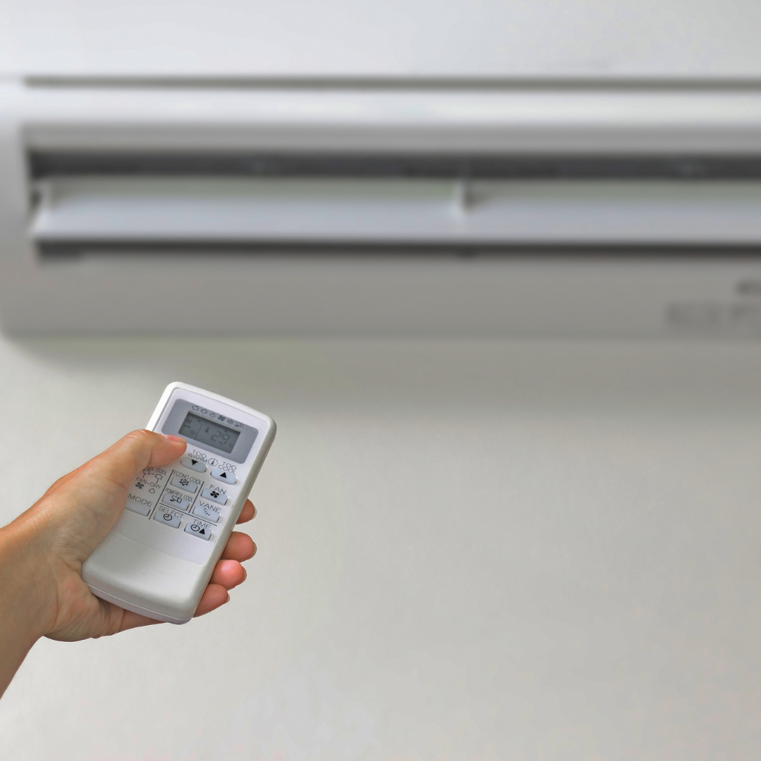 Hand holding remote control, aimed at white air conditioner on a wall.