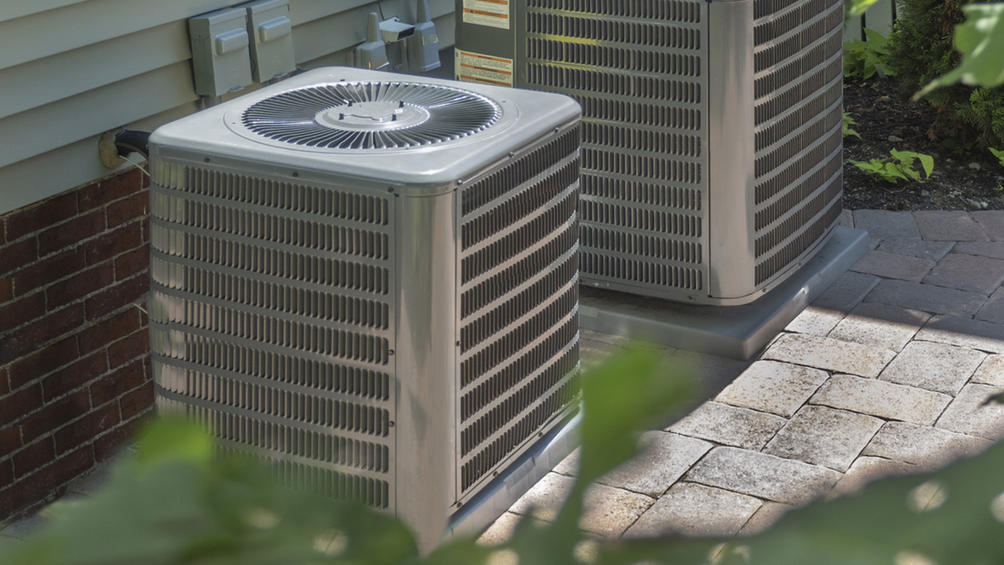 Two outdoor air conditioning units next to a house on a stone patio.
