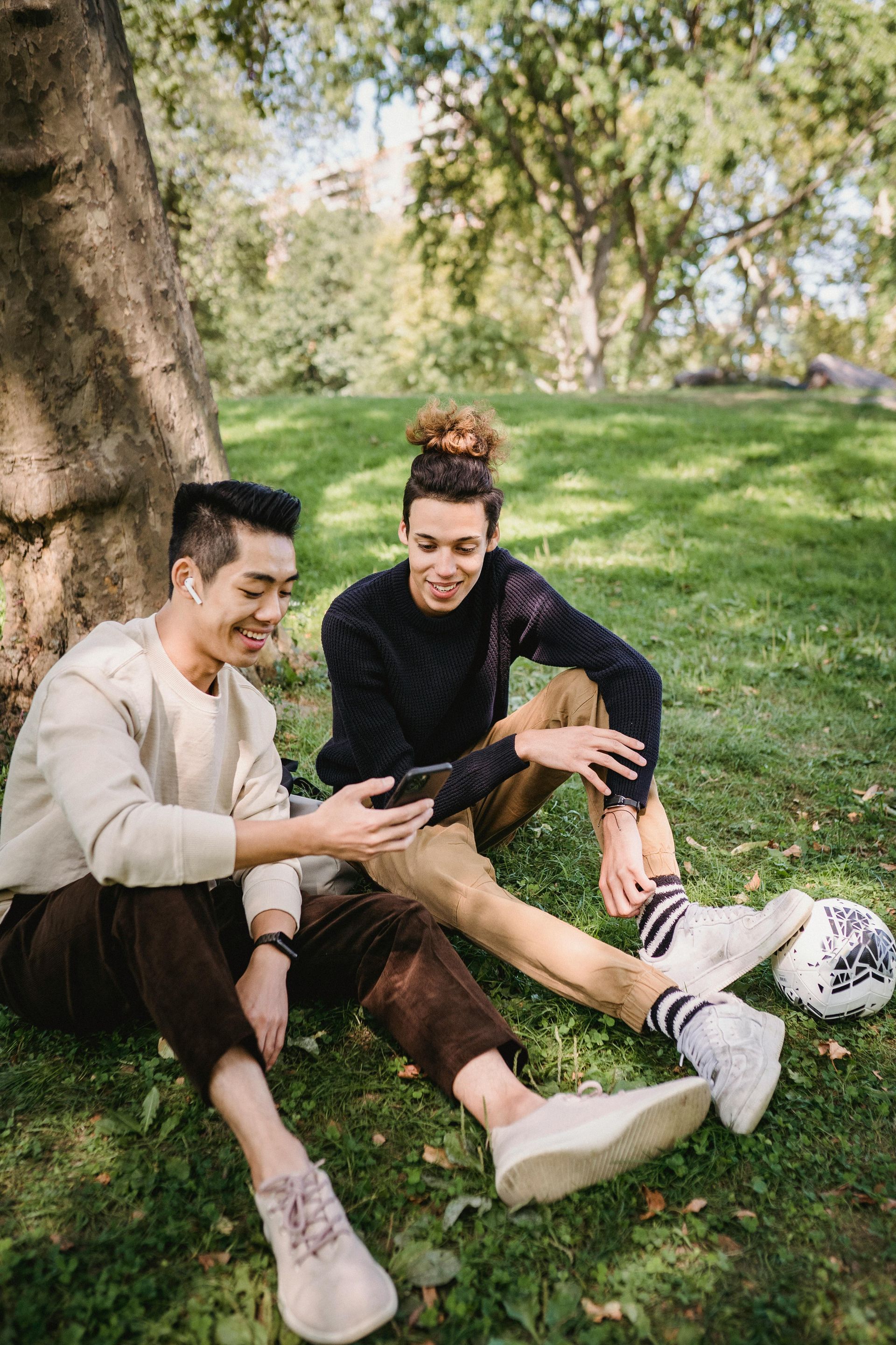 Two men are sitting under a tree in a park looking at a cell phone.