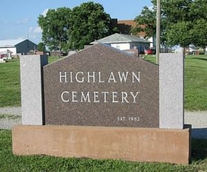 Highlawn Cemetery sign on a stone monument in a grassy outdoor area, with trees and buildings behind it.
