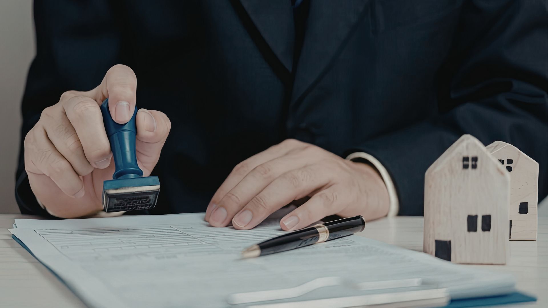 A person in a suit stamps a document next to small wooden house models on a desk.