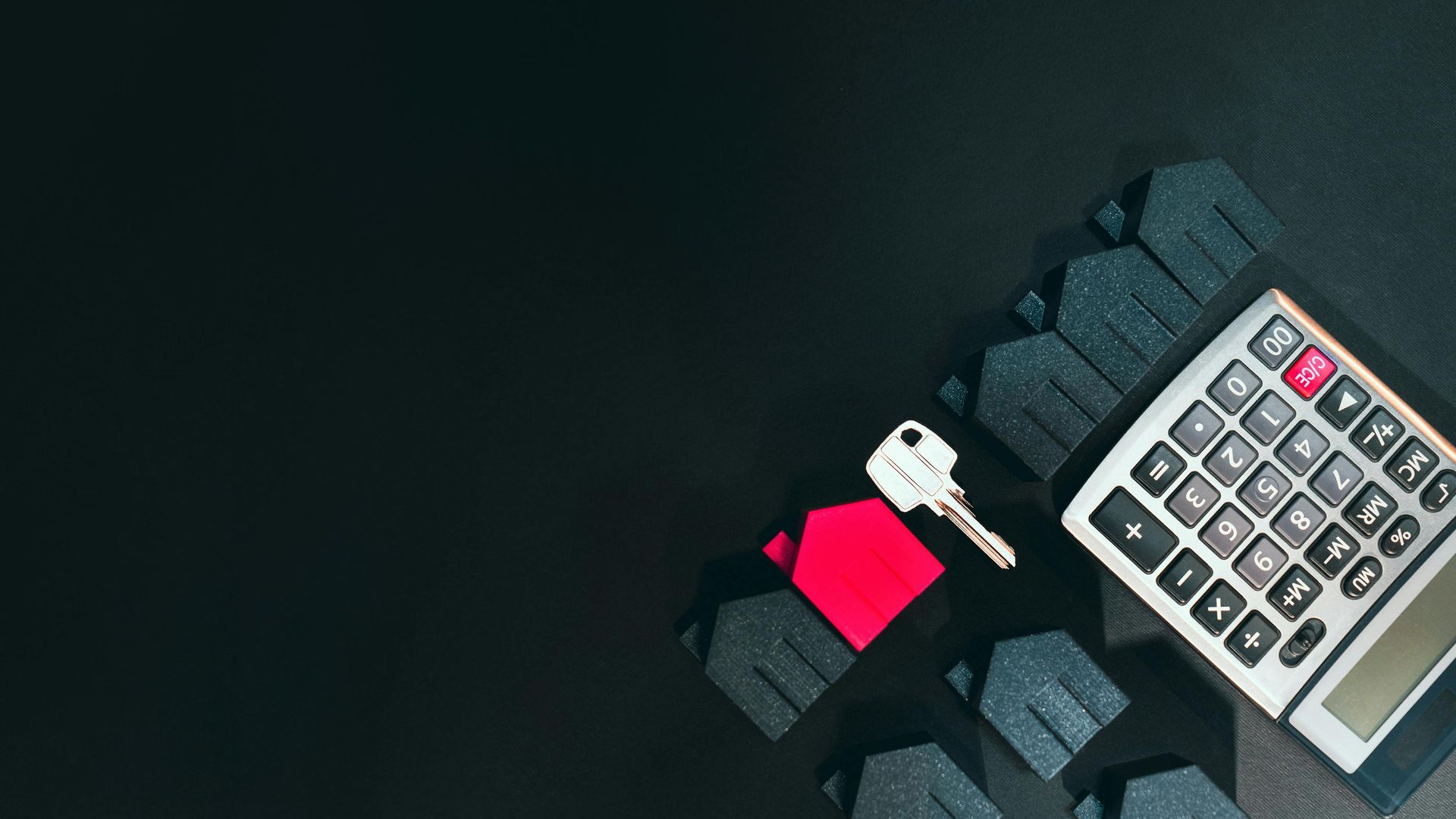 Calculator and key on a black surface next to small house shapes, one red.