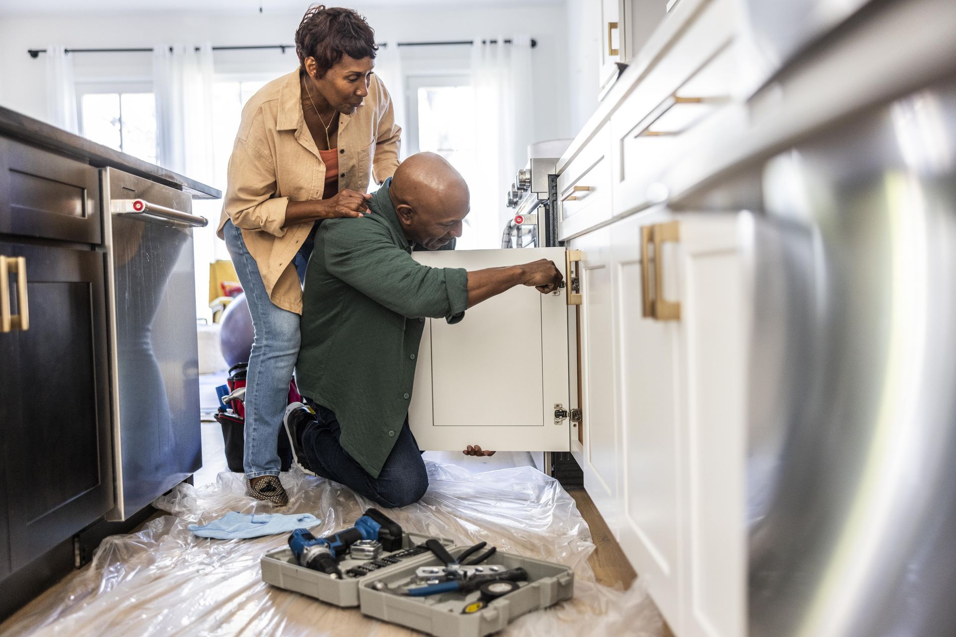 Two people are fixing a kitchen cabinet with tools on the floor.