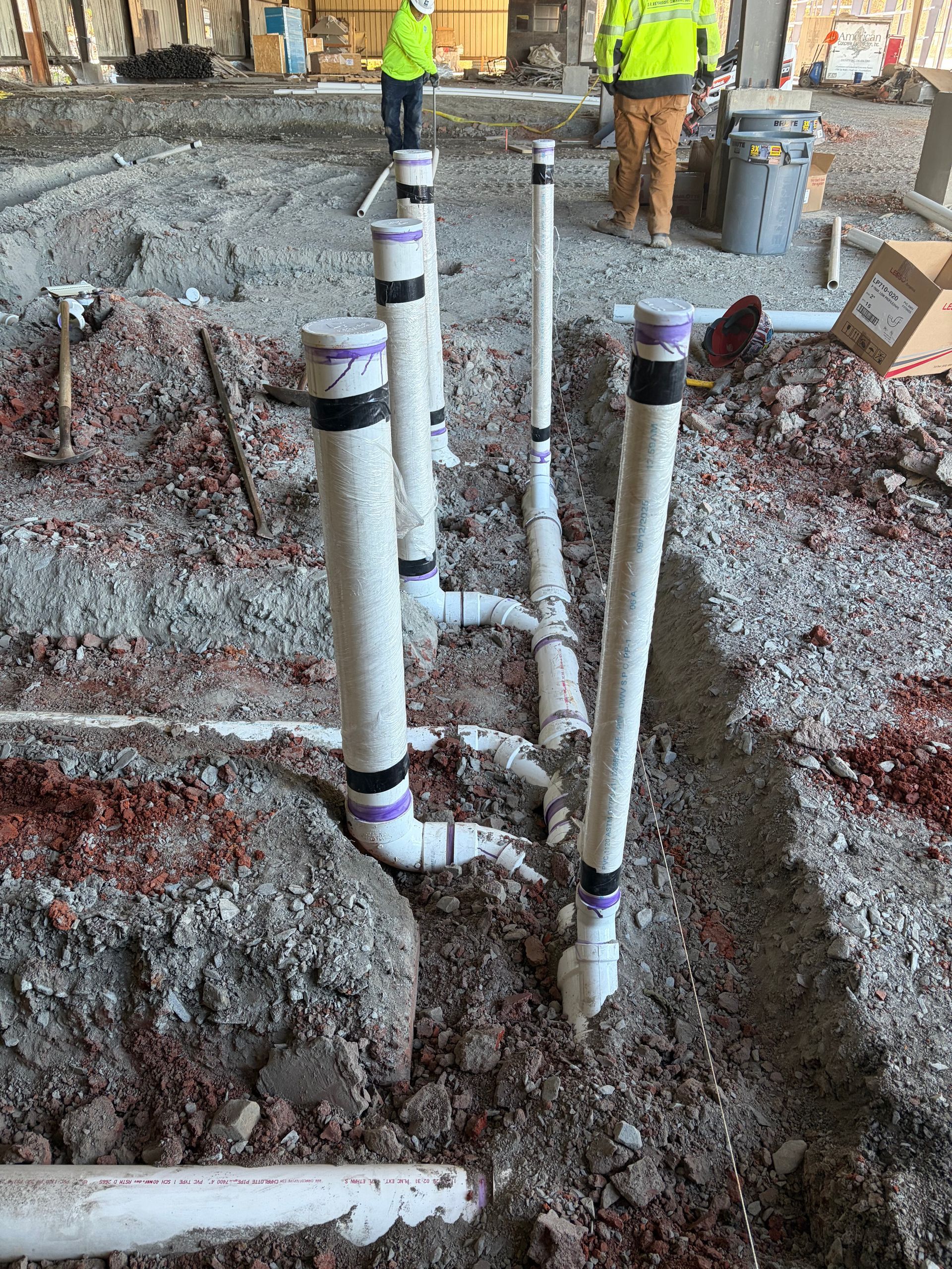 Plumbing pipes installed in a trench on a construction site. White PVC pipes with purple markings; workers in background.