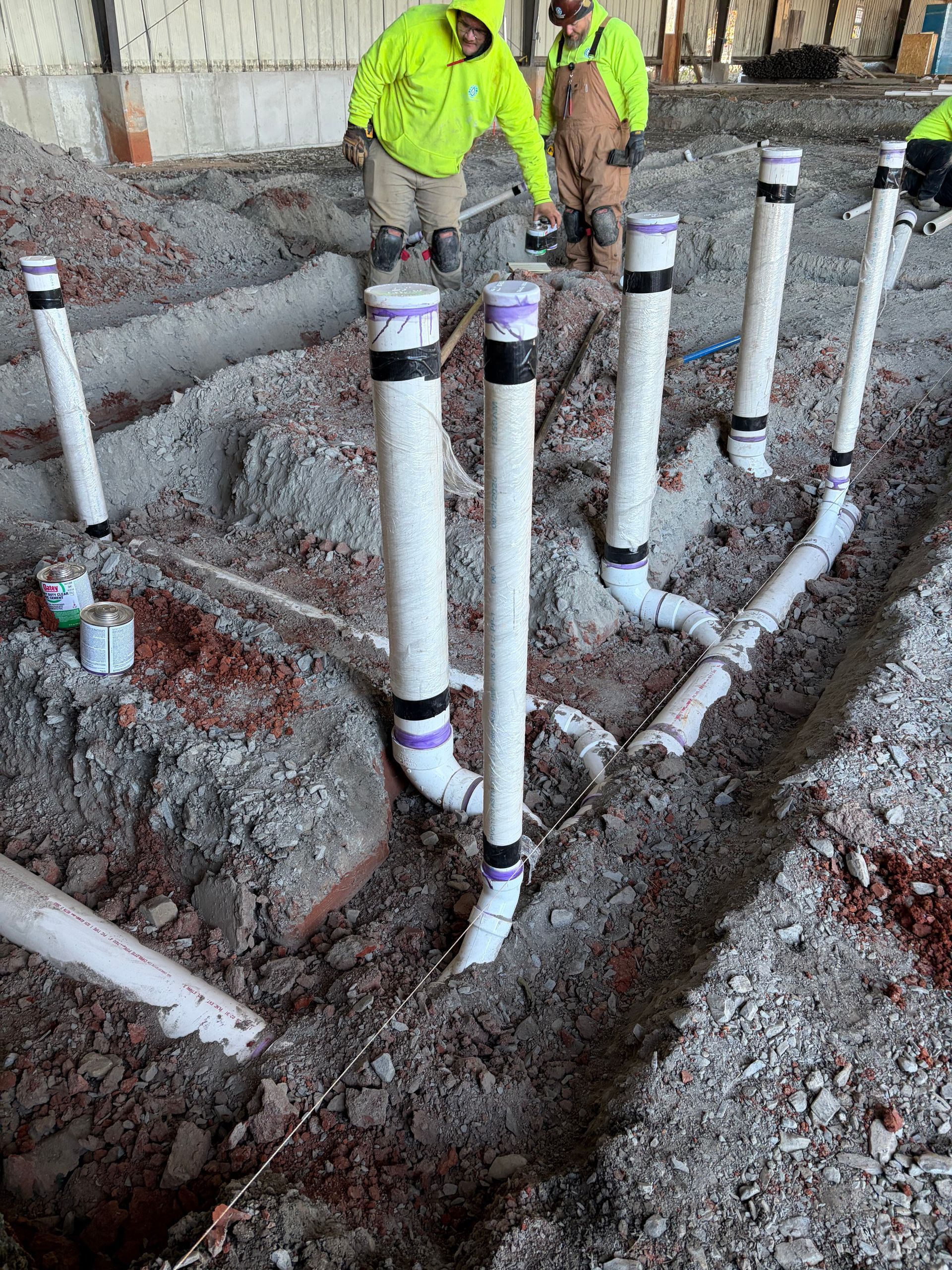 Plumbing installation in construction site. White PVC pipes set in dirt, two workers in safety vests.