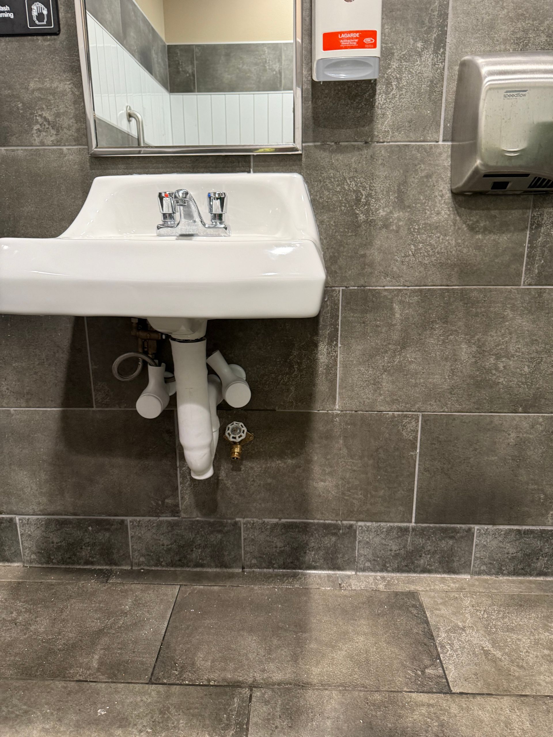 Public restroom sink with faucet, mirror, and hand dryer against gray tiled wall.