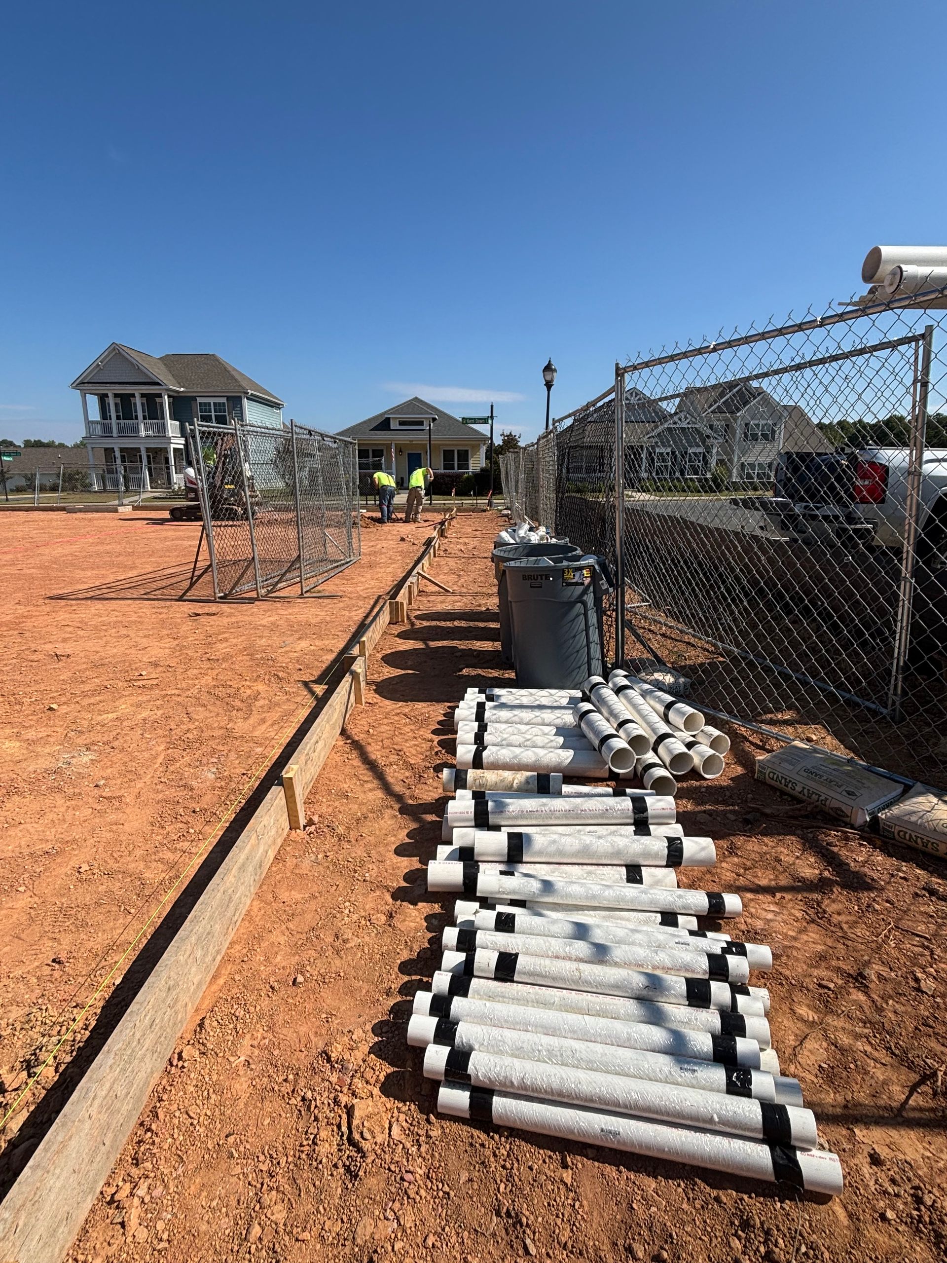 Construction site with white pipes, a chain-link fence, and houses in the background under a blue sky.