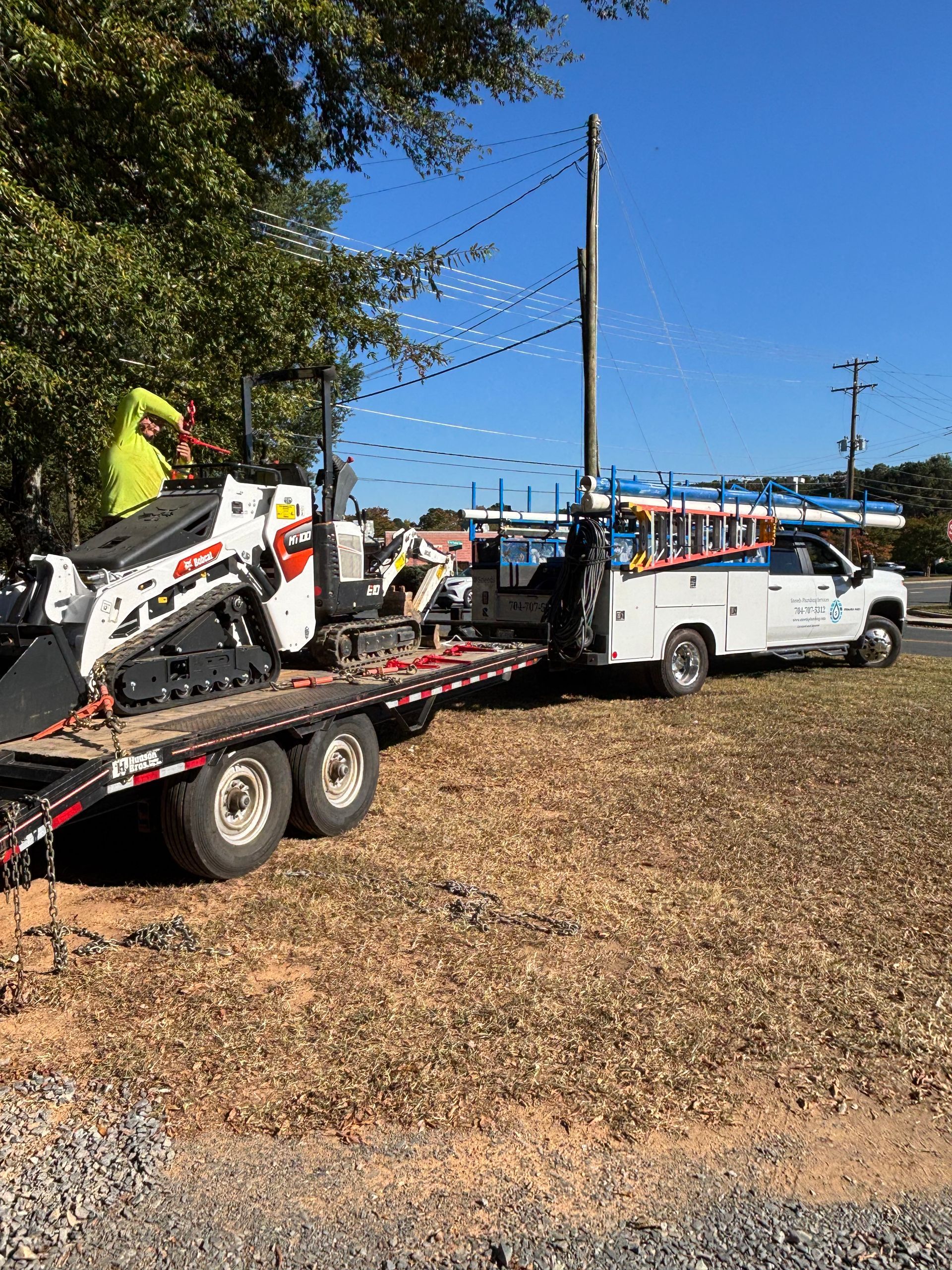 Utility truck with trailer, skid steer, and utility pole. A worker is visible. Sunny day.