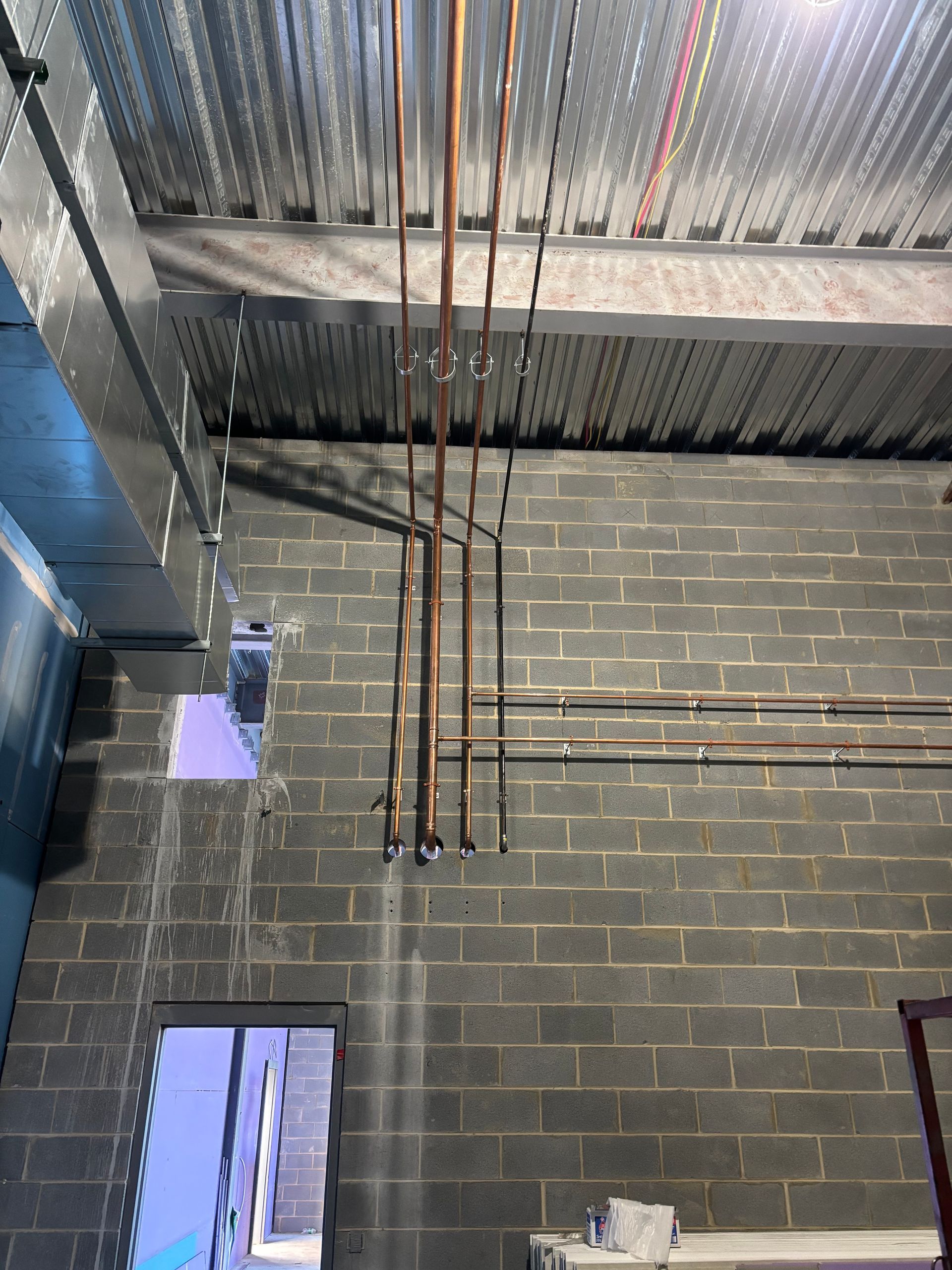 Copper pipes suspended from a corrugated ceiling, against a gray brick wall with an open doorway.
