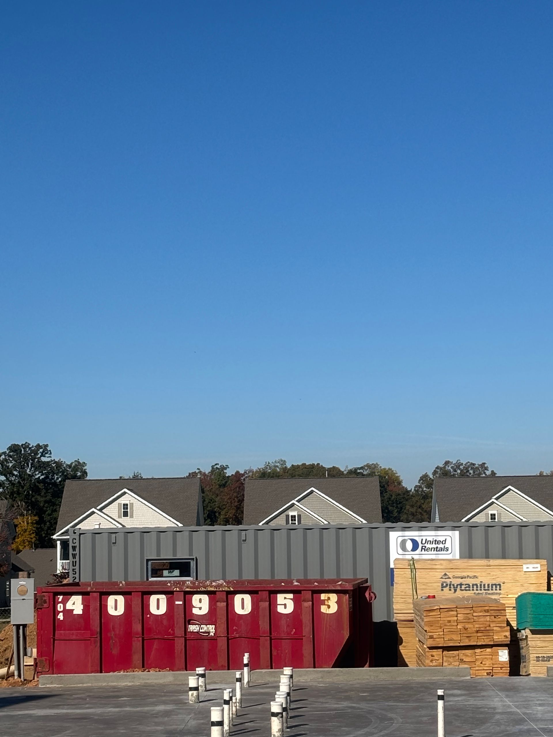 Construction site with a red dumpster, lumber, and houses against a blue sky.