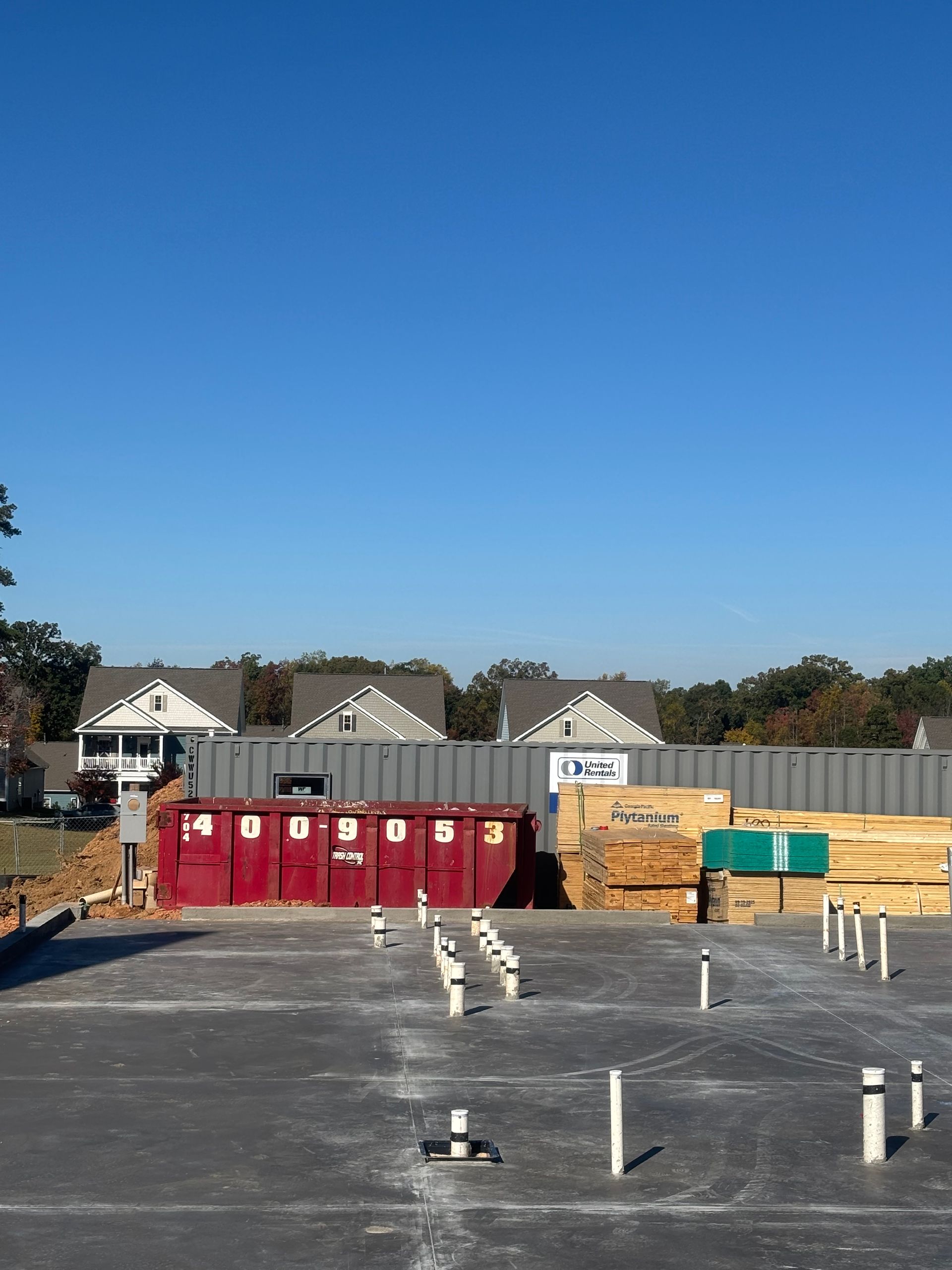 Construction site with a red dumpster, lumber, and houses in the background under a blue sky.