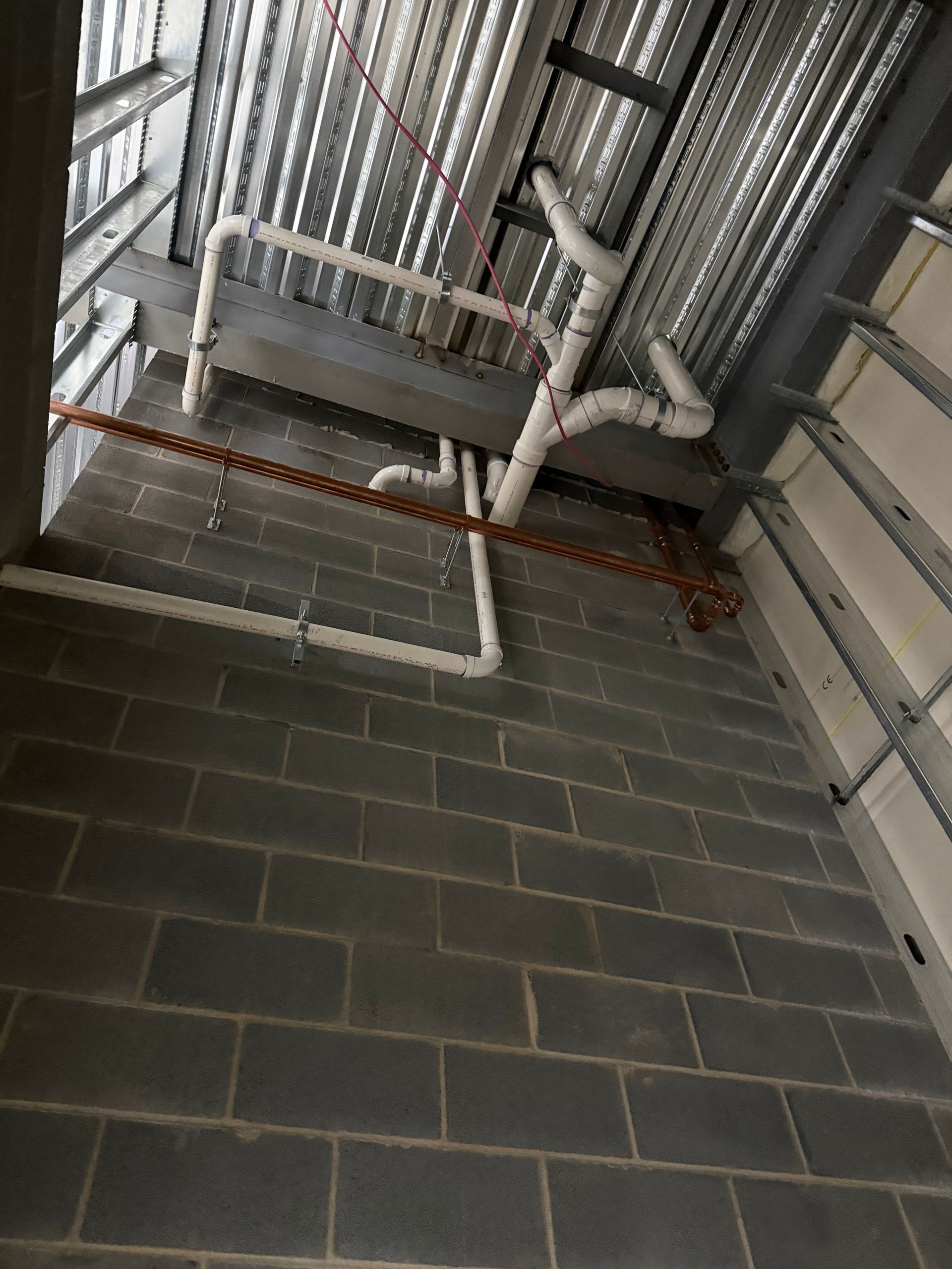 Plumbing pipes and conduit installed in a construction setting with gray concrete block wall and metal ceiling.