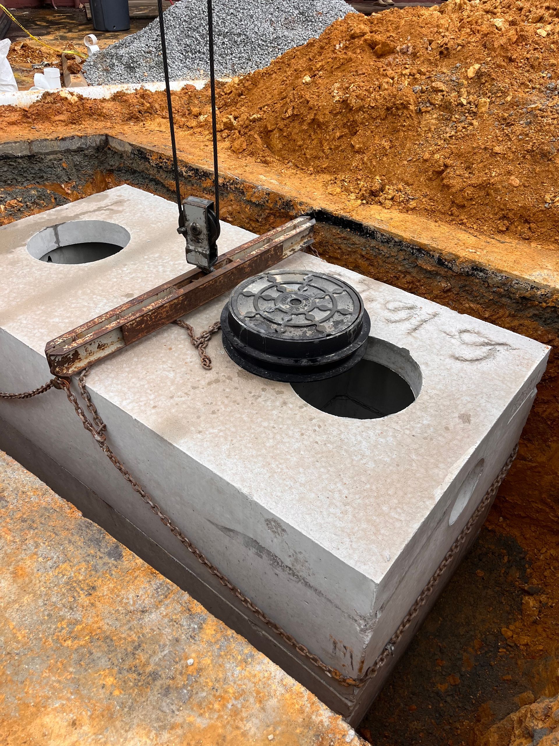Concrete septic tank being installed in a dirt pit, with chains and access hatch open.