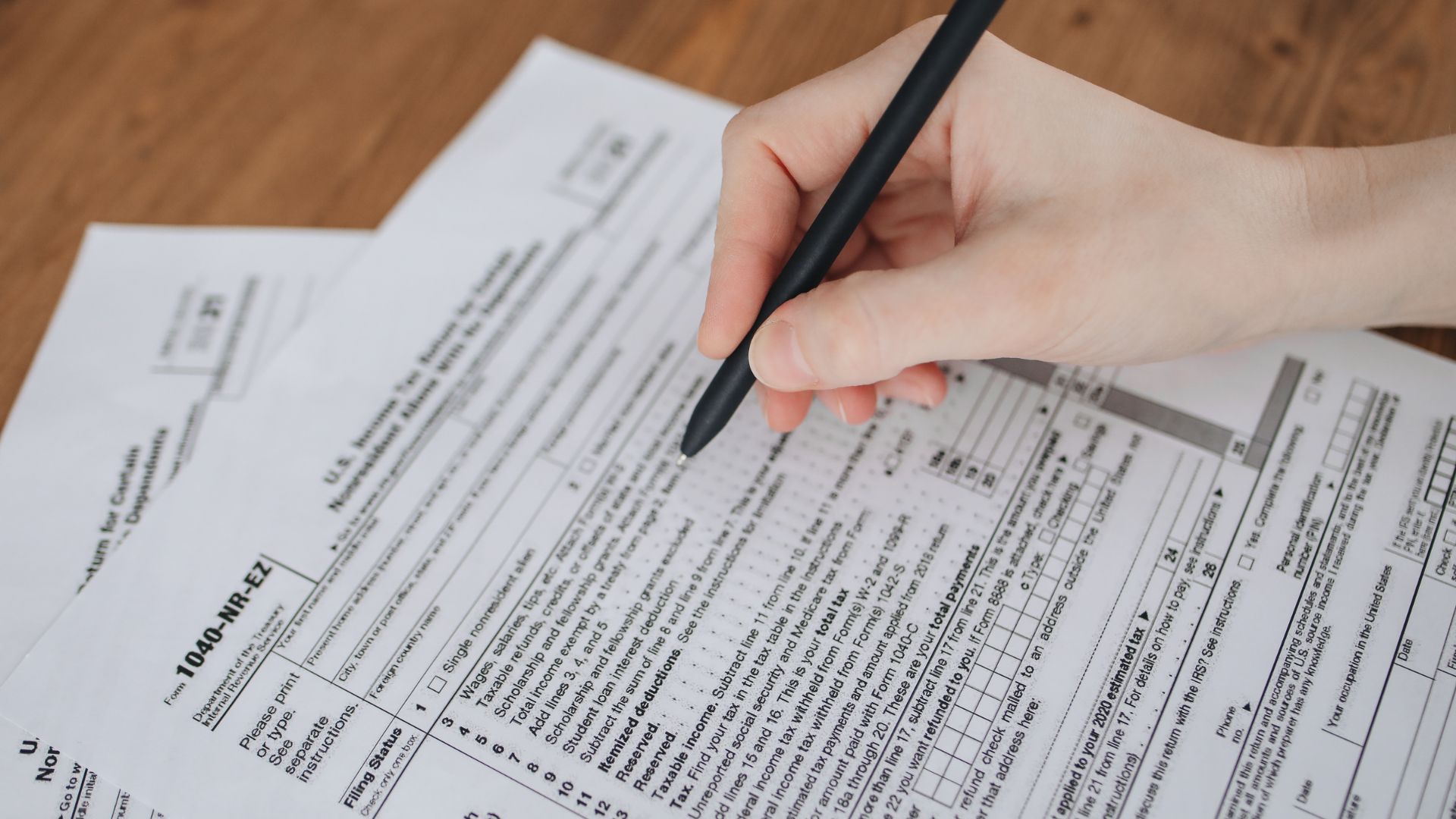 Hand writing on tax forms with a black pen on a wooden table.