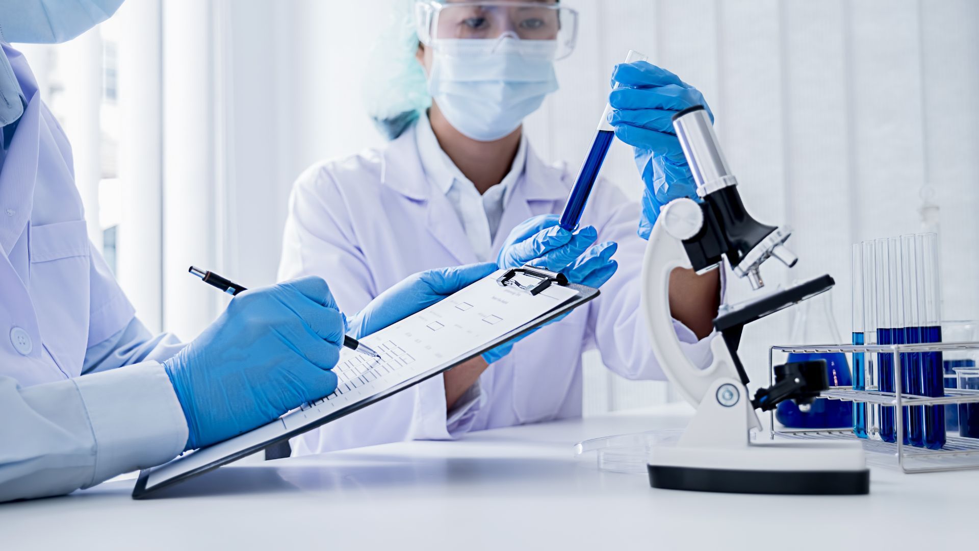 Two lab workers in protective gear examine a test tube, writing notes near a microscope.