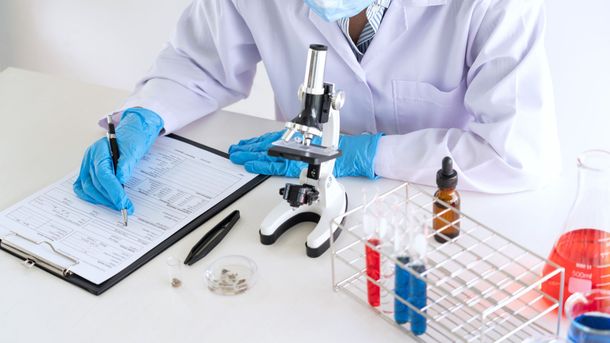 Scientist in lab coat and gloves writes notes, next to microscope, test tubes, and chemicals.