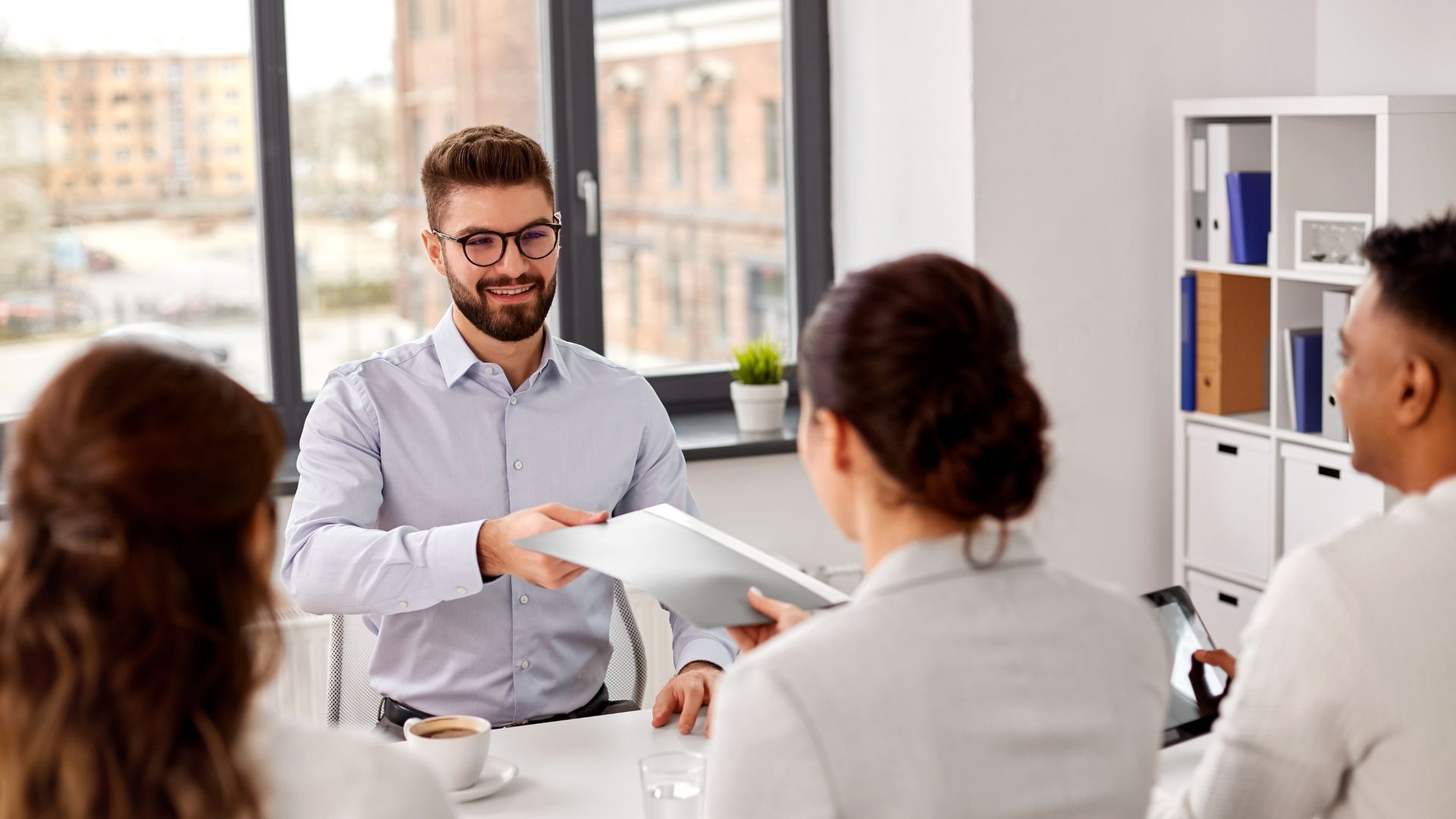 Man in a light blue shirt hands papers to a woman at a table during a meeting.