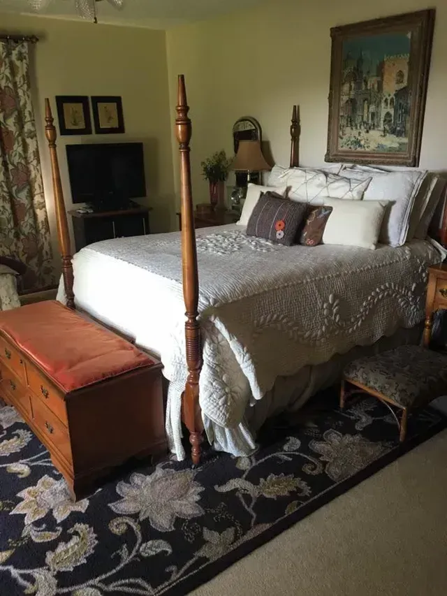 Bedroom with a four-poster bed, floral rug, wooden chest, and framed artwork