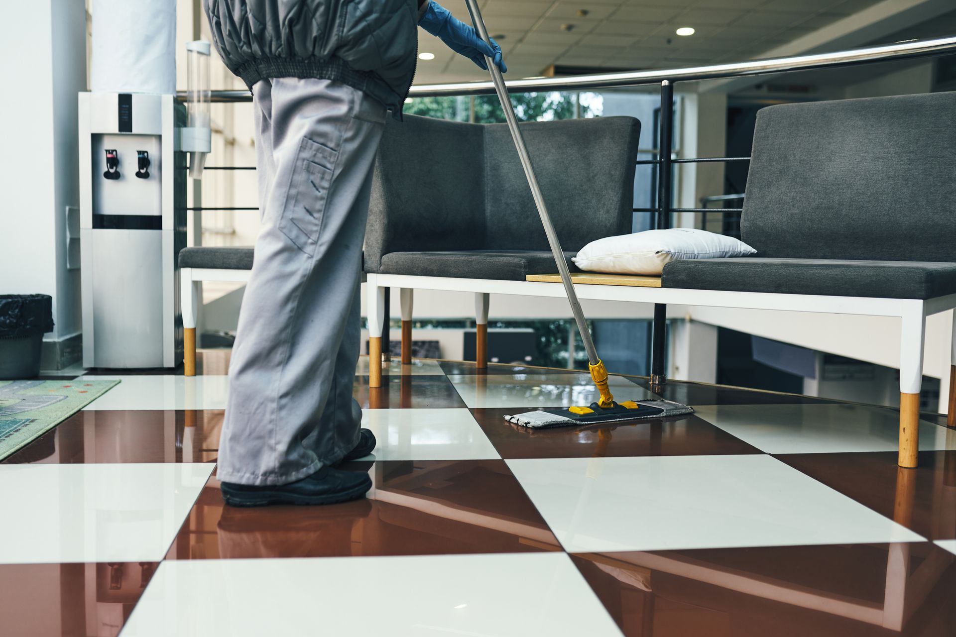 A janitor mopping a checkered tile floor in a modern lobby area with chairs and cleaning tools
