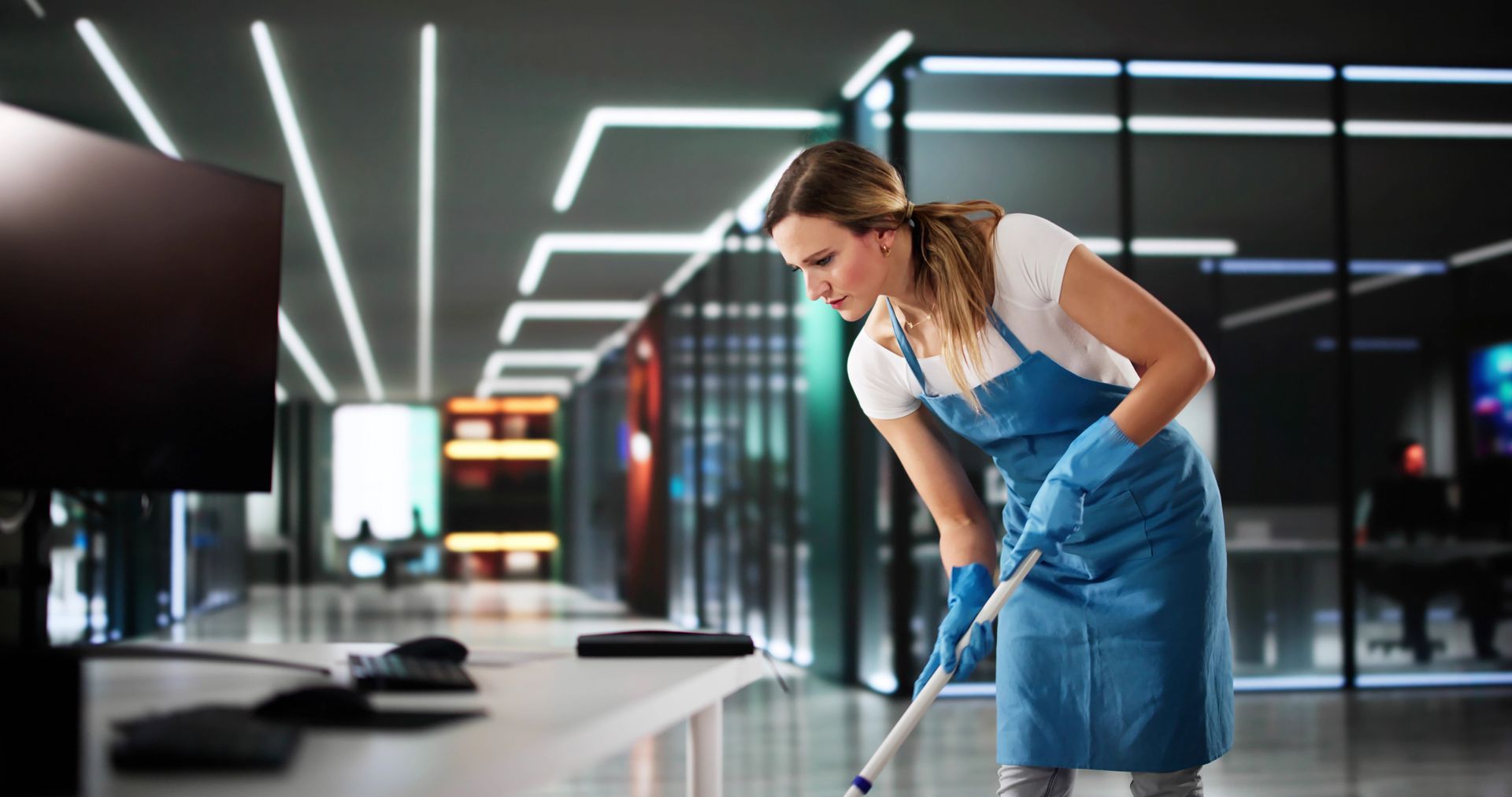 A cleaner wearing a blue apron wipes a desk surface in a modern office with glass partitions.
