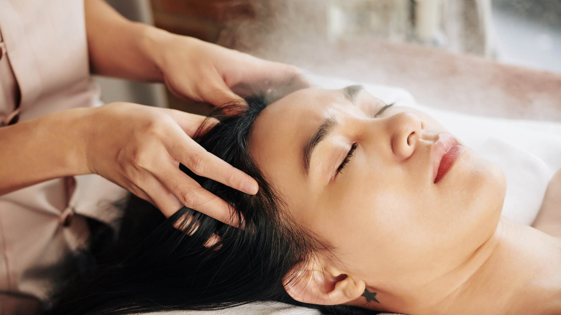 Woman receiving a scalp massage with mist; eyes closed, spa setting.