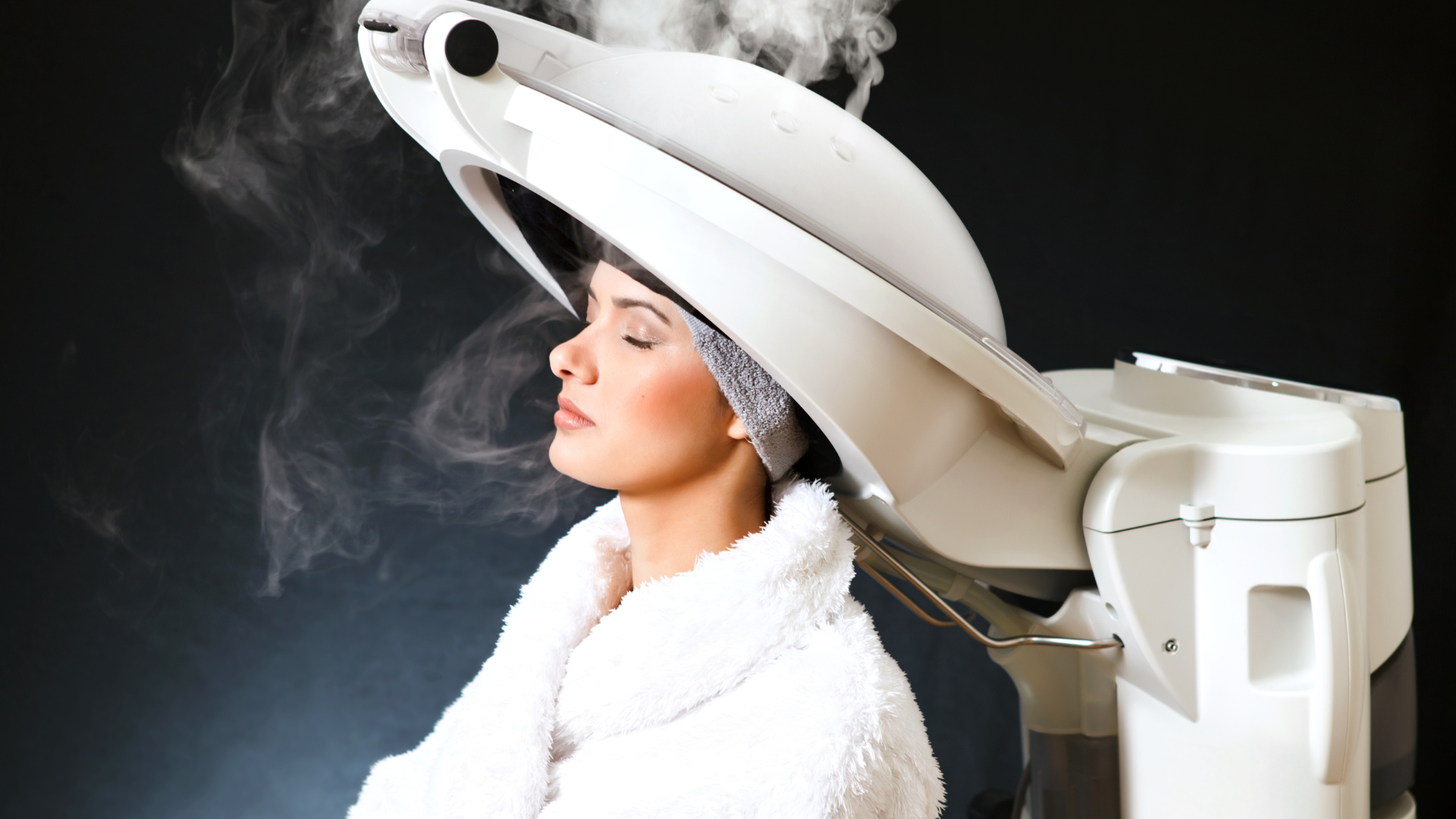 Woman getting a hair steam treatment at a salon, wearing a white robe, eyes closed, against a dark background.