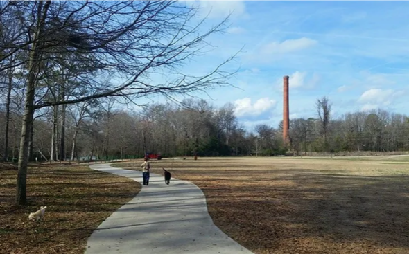 A person walking a dog down a path in a park with a chimney in the background.