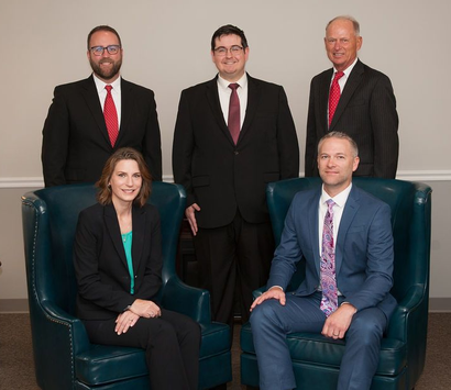 Three people in suits stand behind two others seated in teal armchairs in an office setting.