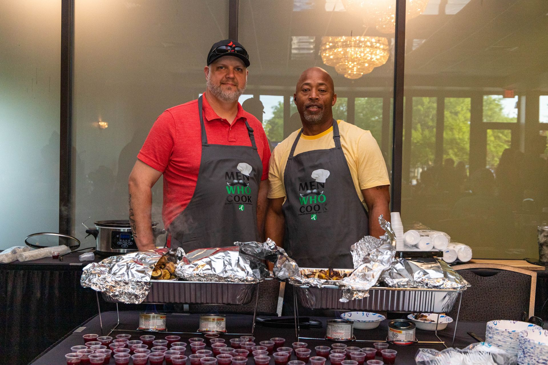 two men wearing aprons are standing next to each other in front of a table filled with food .