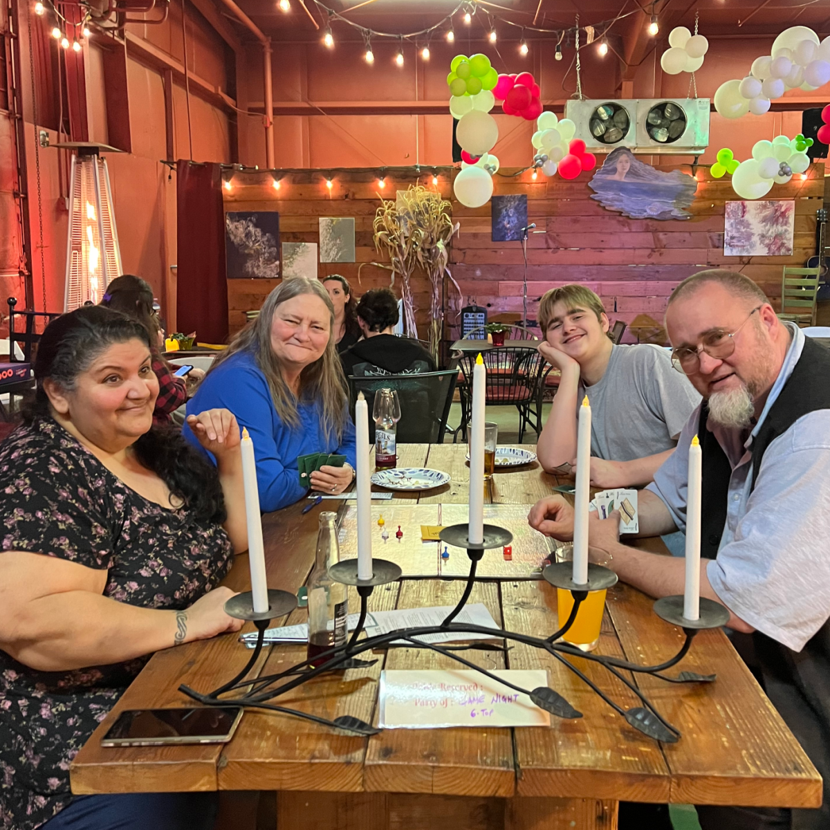 Four people, of three generations, sitting at the big wood plank table where they have been playing a variety of board games, smiling at the camera.