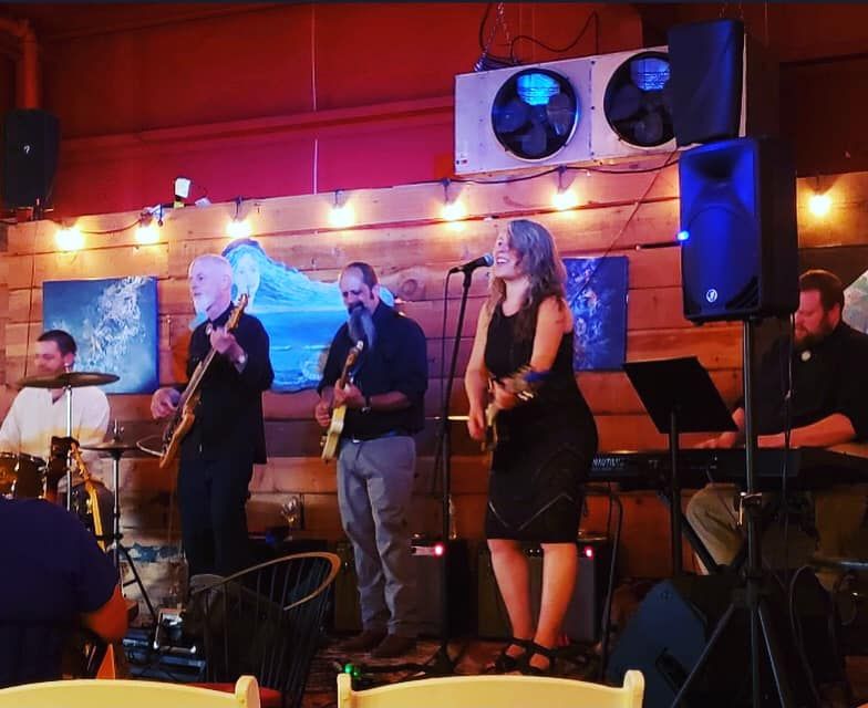 Three musicians playing on the Oran Mor stage with lights above their heads and the wood plank backdrop with the paintings on it.