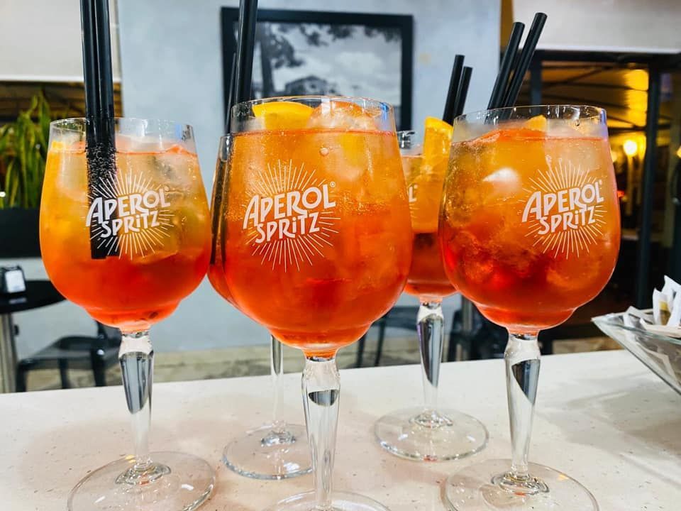 Quattro cocktail Aperol Spritz con ghiaccio e fette d'arancia, serviti in bicchieri personalizzati su un bancone del bar.