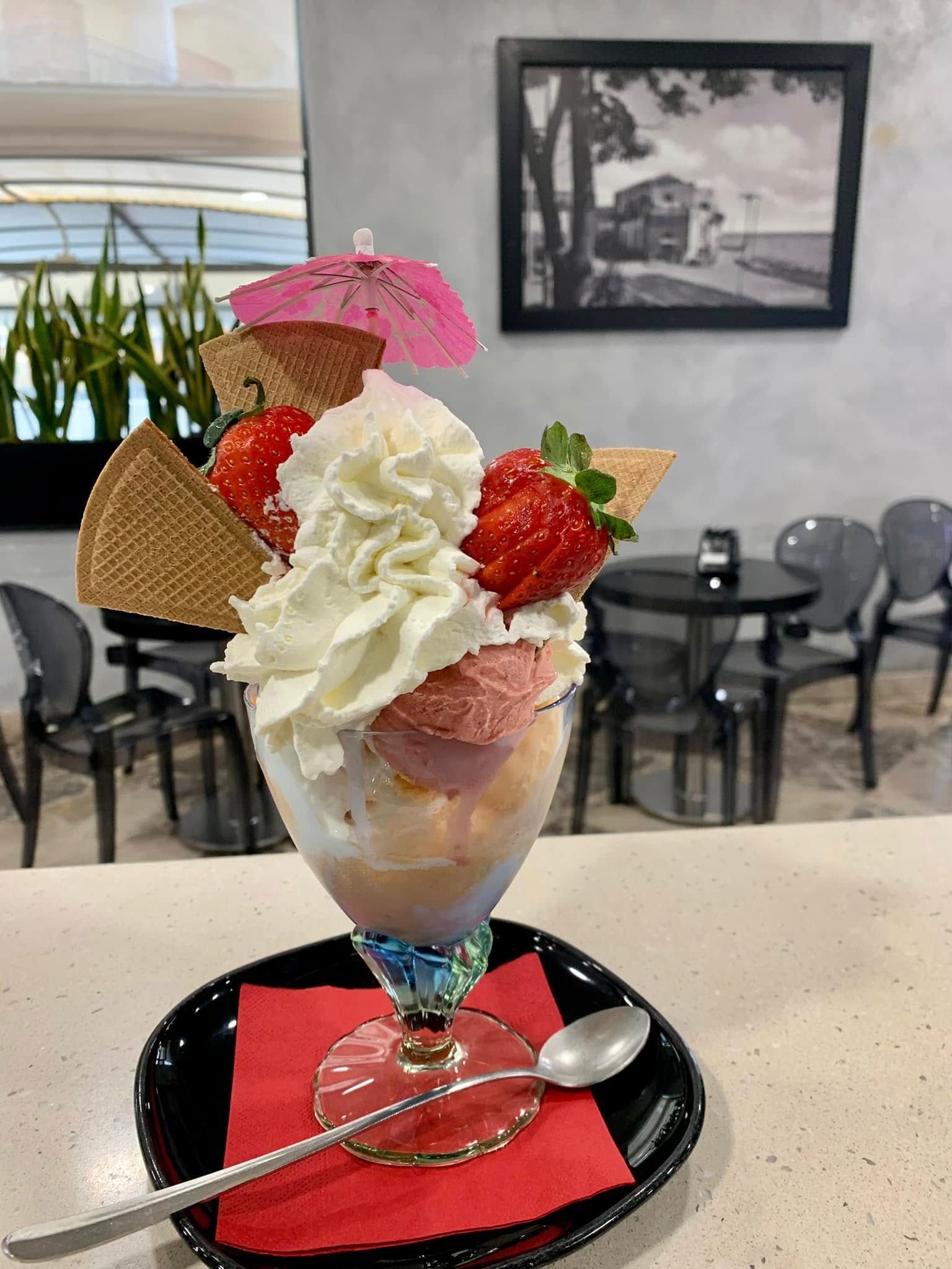 Una coppa gelato decorativa con panna montata, fragole e un ombrellino rosa in una coppetta di vetro su un tavolino.