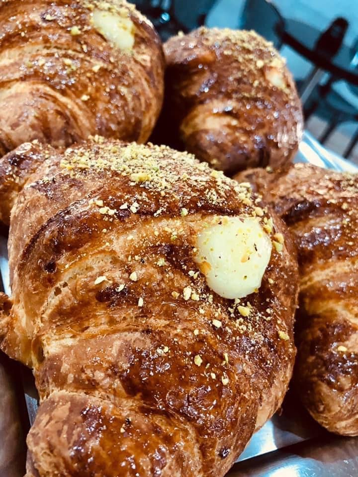 Diversi croissant dorati, cosparsi di granella di frutta secca e ripieni di crema, disposti su un vassoio di metallo.