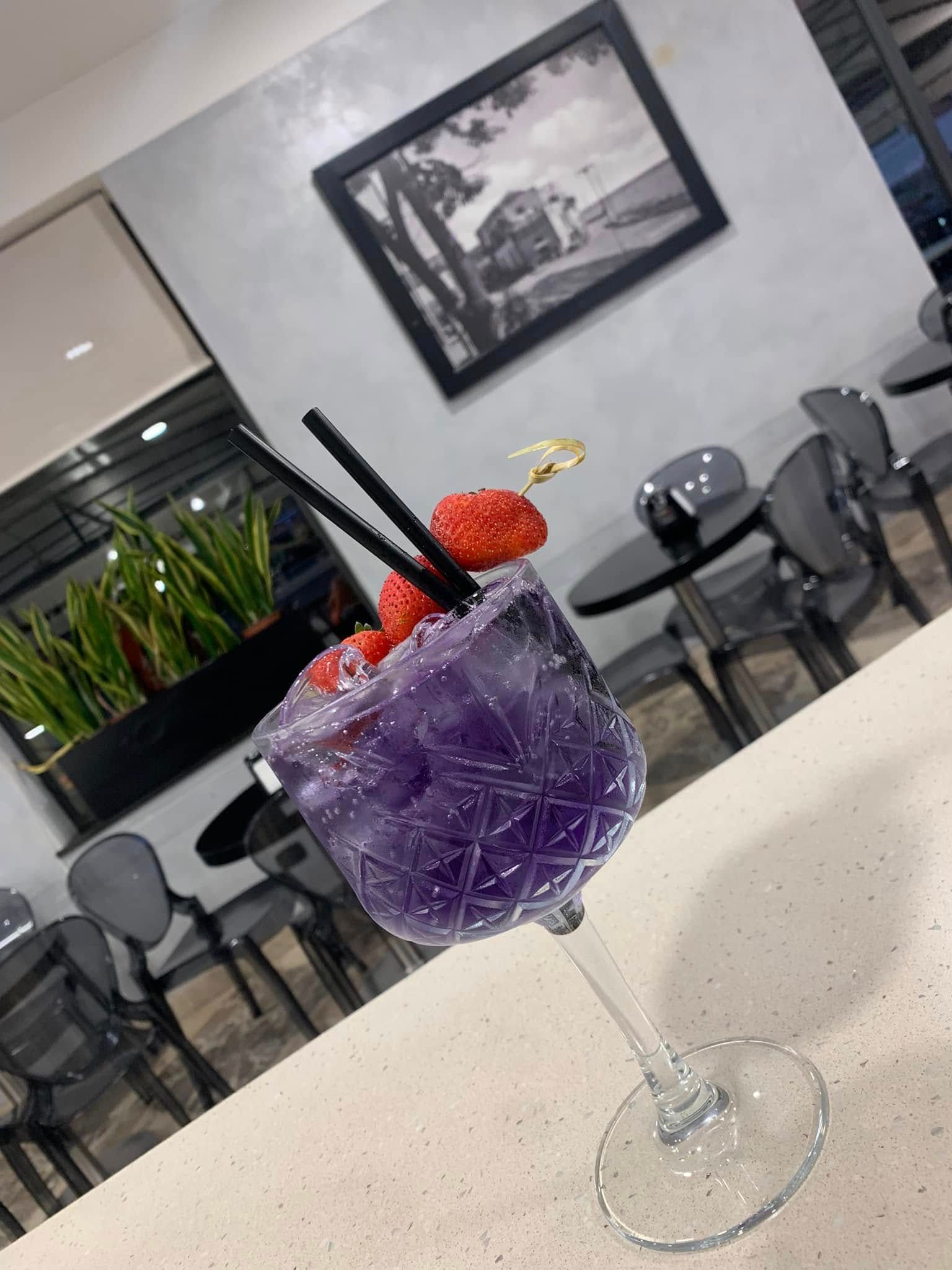 Un cocktail viola in un bicchiere dalla superficie ruvida, guarnito con una fragola, appoggiato su un bancone in un ristorante moderno e minimalista.