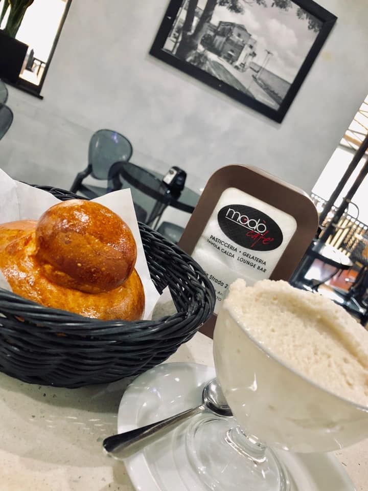 Al Modo Cafe, un panino brioche in un cestino nero è appoggiato accanto a un bicchiere di granita di mandorle su un tavolino.