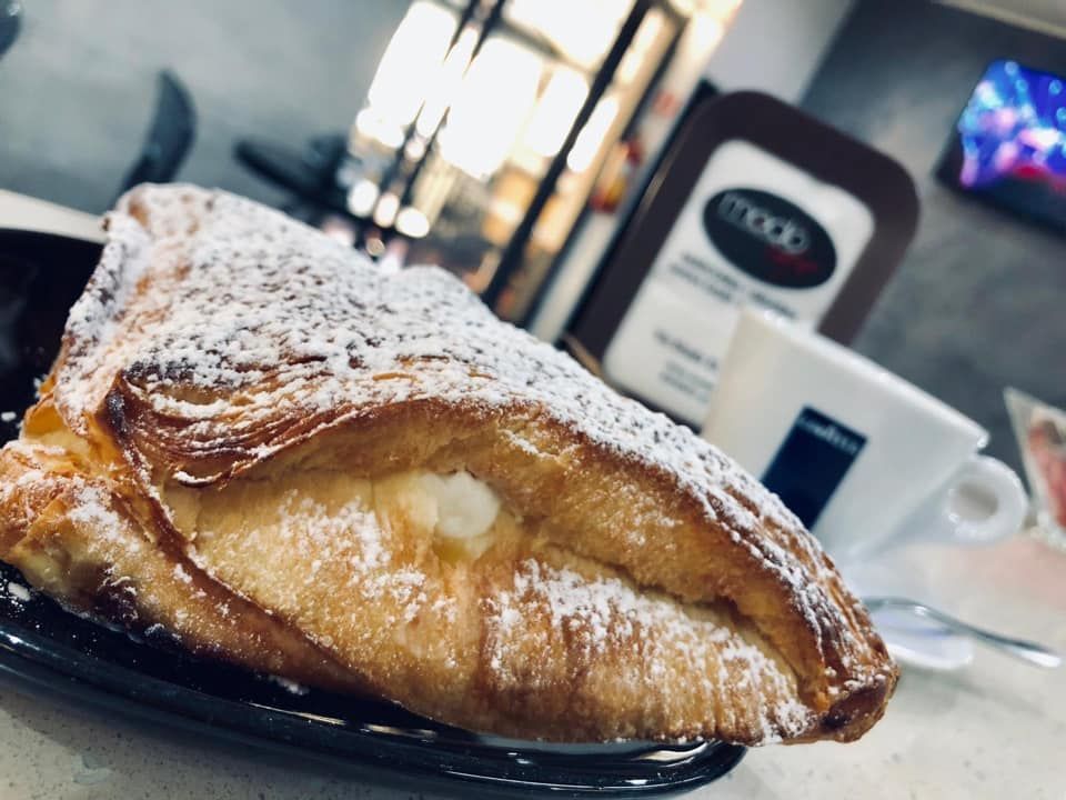 Un pasticcino ripieno e cosparso di zucchero a velo, servito su un piatto nero con una tazza di caffè bianca, in un'atmosfera da caffetteria.