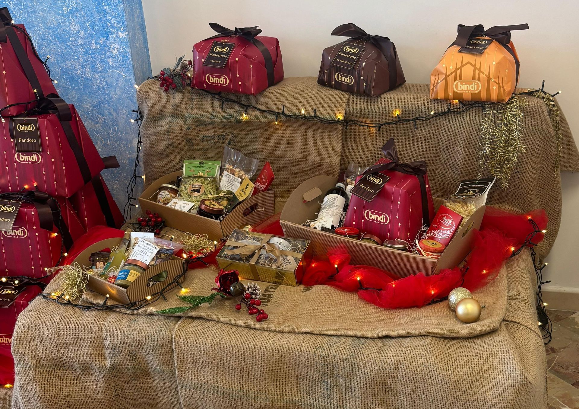 Cesti regalo e panettoni incartati, esposti su un tavolo ricoperto di tela di iuta, illuminato da lucine e addobbi festivi.