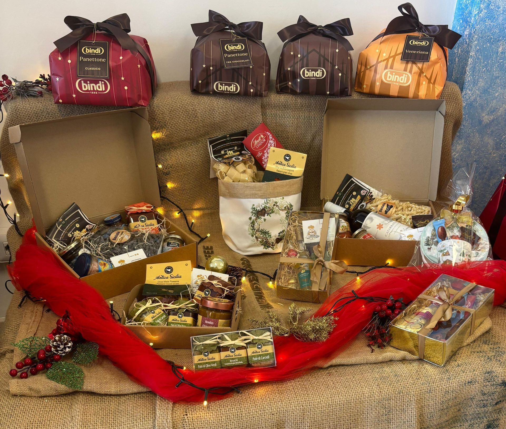 Cesti regalo natalizi con panettone incartato, scatole festive e vino, disposti su una superficie di iuta con luci decorative.