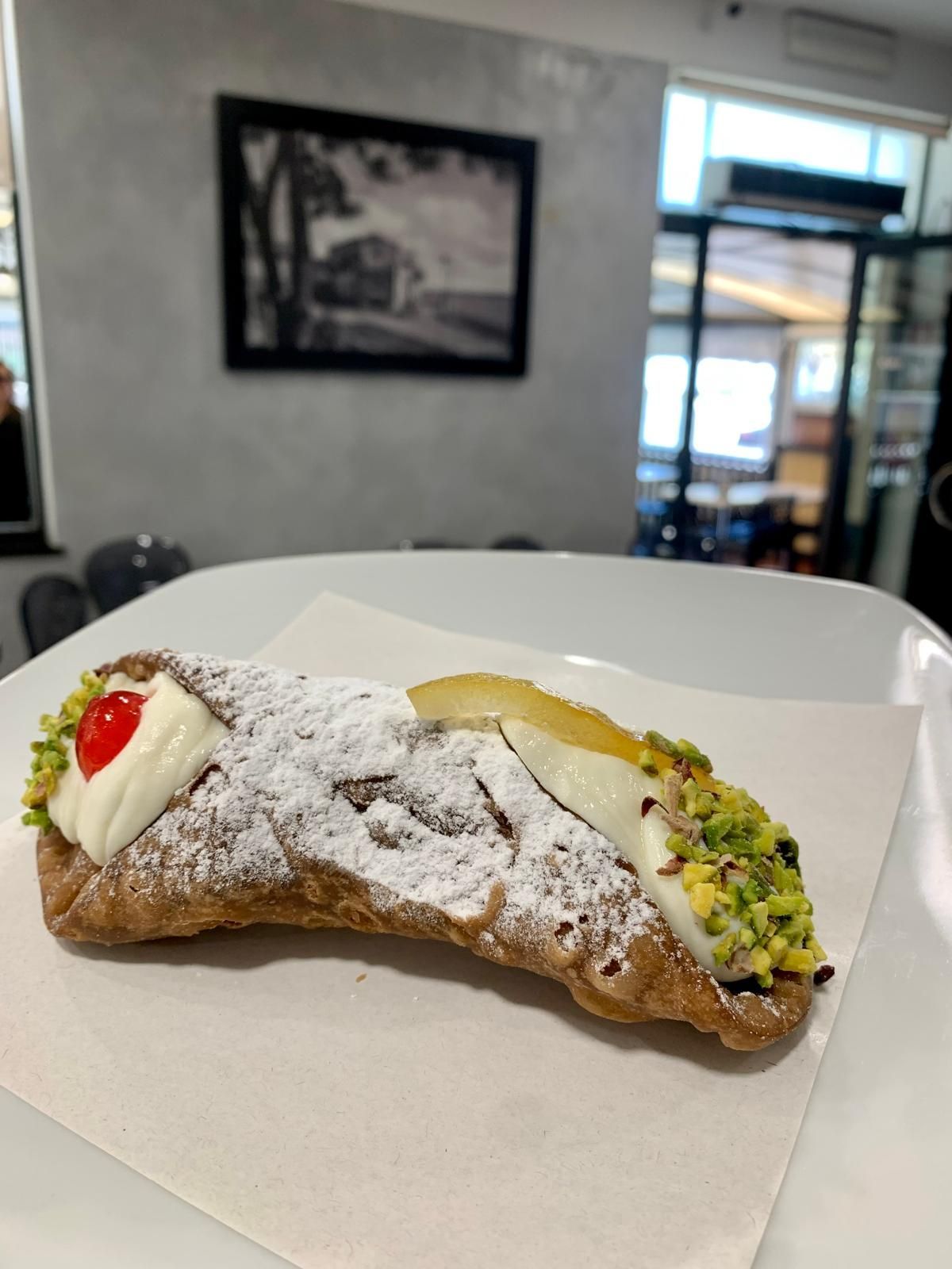 Una persona tiene in mano un cannolo ricoperto di pistacchio e spolverato di zucchero a velo, con sullo sfondo un'insegna sfocata del 