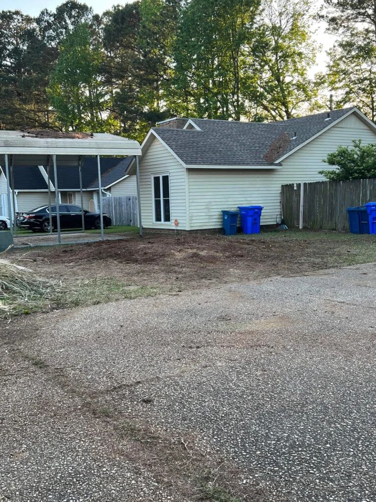 A white house with a carport and blue trash cans in front of it.